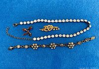 Three costume jewelry pieces on blue background: silver-tone rhinestone bracelet with floral motifs, a delicate rhinestone chain bracelet or necklace, and a gold-tone heart and arrow brooch with faux pearls.