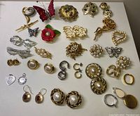 Full display of assorted costume jewelry laid out, showing brooches shaped like flowers, butterflies, leaves, hearts and assorted earrings including clip-on and pierced with faux pearls and colorful stones