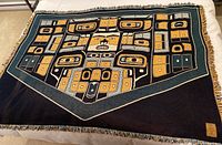Full view of The Chilkat patterned blanket showing the intricate Indigenous design in blue, mustard yellow, teal, and white with fringed edges.