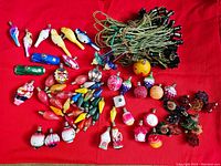 Wide shot showing a large assortment of 69 assorted vintage Christmas bulbs and three tangled green cord light strands on a red surface, including animal, bird, fruit, flower, clock, and face designs.