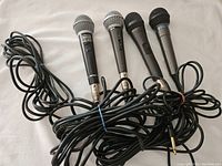 Four wired microphones laid side by side, cables coiled and tied with rubber bands, showing metal grille and black cylindrical body. Visible switches on each microphone.