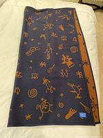 Folded Pendleton blanket showing dark blue side with rust orange primitive motifs laid flat on a bed.