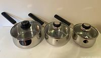 Three Lagostina stainless steel pots with glass lids positioned side by side showing different sizes and black handles.