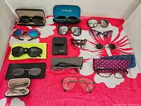 Image showing an assortment of 14 pairs of sunglasses and readers arranged on a red and white patterned cloth, including several cases and sleeves.