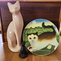 Front view showing pink ceramic cat statue, small dark carved cat figurine, and colorful Martin Lehman cat plate with scenic background.