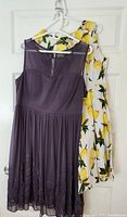 Two dresses hanging on white hangers on a door: purple sleeveless dress with sheer upper part and floral hem, white sleeveless lemon print dress