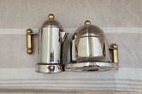 Side view of paired stainless steel cream pitcher and sugar bowl with gold plated handles and lid knobs on a beige striped fabric background.