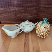 Photo showing white porcelain sugar bowl with lid and matching creamer, both decorated with gold leaf accents, alongside the pineapple-shaped lidded sugar bowl.