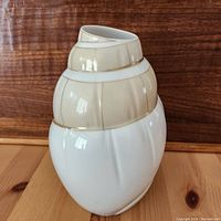 Side view of spiral seashell shaped vase with cream, white, and gold detailing on wooden tabletop and background