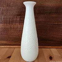 Front view of tall white bisque vase with embossed floral design on wooden surface against wooden backdrop.