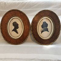 Two matching vintage oval wooden frames with gold accents containing black needlework silhouette portraits on beige fabric backgrounds, profiles of a woman and a man.