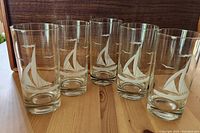 Front view of all five clear glass tumblers with etched sailboat and seagulls design, arranged in a row on a wooden surface with a wooden background.