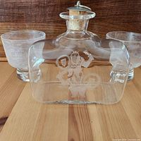 Front view of Viking-themed clear glass decanter with silver plated cork top, showing etched Viking design and two matching glasses behind it.