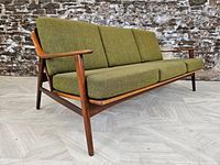 Front perspective of three-seat teak sofa with olive green cushions, angled against a stone wall.