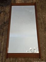 Full view of rectangular mirror with teak wood frame against wood floor.