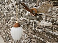 Full view of wall sconce mounted on textured stone wall, showing wooden bracket, brass rod, cord, and glass shade.