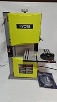 Front view of Ryobi tabletop bandsaw on white background, showing power switch, work table with miter gauge, and spare blade package