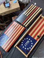 Three handcrafted wooden flags laid flat: one traditional 1776 design, one firefighter flag with black and red stripes, and one 1776 design with hooks and framed edges.