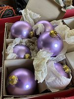 Close-up of multiple lavender colored Christmas ball ornaments inside compartmentalized sections of original white cardboard box with tissue paper protection.