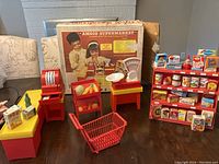 Overall view of supermarket playset including original box, cash register, plastic produce bin with fruit, weighing scale on table, plastic conveyor shelf with miniature groceries, and red shopping cart missing wheels.