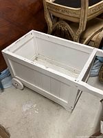 White wooden cart with rectangular box shape, front wheels, and handle on floor near chairs.