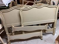 Two twin headboards and two footboards stacked upright showing cream color with gold accents and floral carved details.