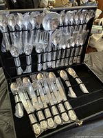Silverware pieces arranged inside carrying case, wrapped in plastic, with ornate decorative handles visible. Forks, knives, spoons and serving utensils shown.