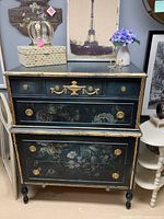 Front view of navy blue dresser showing all six drawers, floral transfer decorations, gold leaf trim and hardware. Items on top include decorative box, crown figurine, vase with flowers, and framed artwork.