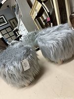 Pair of gray faux fur footstools shown from an angle, displaying fluffy texture and shape.