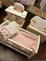 Photo showing white wooden crib bed with fabric accessories including stuffed mattress, floral and pink blankets, pillow, and pillowcase. Also shows bouncy chair and rocking chair with boxes in background.