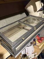 Photo of the rectangular wooden and glass display case resting on a shelf, showing the metallic gray paint and glass top divided into two sections, hinge and latch visible.