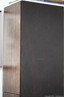 Side view of one speaker showing wood veneer cabinet and black fabric grille cover.