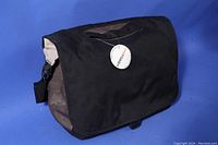 Black over-shoulder camera bag with flap closure and side pouches against blue background.