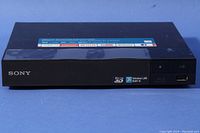 Front view of Sony Blu-Ray player showing power button, disc tray, USB port, and branding.