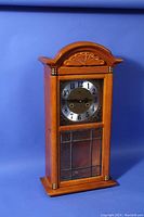 Front view of wooden clock case with carved arched top, metal dial with black numerals, and leaded glass lower panel under dial.