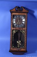 Front view of vintage wooden pendulum wall clock with ornate top molding, Roman numeral dial, and decorative lower glass panel showing pendulum.
