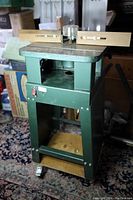 Front view of green metal wood shaper mounted on base with caster wheels, showing flat work surface, adjustable fences, and control switch.