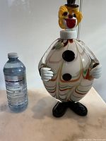 Front view of large hand-blown Murano glass clown decanter next to water bottle for scale showing colorful swirling pattern, black feet base, and clown head stopper with top hat.