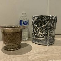Photo showing two vintage planters with a water bottle behind for scale: one round metal intricately engraved planter and one square textured black and white ceramic planter.