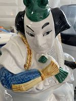 Close-up of the Kabuki Mikado figurine's face and upper body showing detailed facial paint, green headpiece, and colorful sleeves.