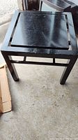 Photo of the black wooden square table on a concrete floor showing top and legs with some scratches and wear.