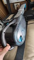 Photo of a black and gray cordless handheld vacuum cleaner held in hand, showing charging port and dust container with ergonomic handle.