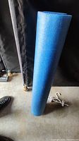 Photo showing a large cylindrical blue foam exercise roller standing upright on a concrete floor in front of a dark backdrop.