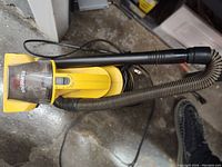 Yellow Eureka handheld vacuum cleaner on concrete floor with attached flexible hose and black extension wand visible from top angle