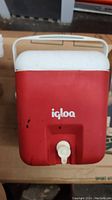 Front view of red Igloo water dispenser with white lid and handle, showing black markings on red body and white dispensing spigot on front bottom center.
