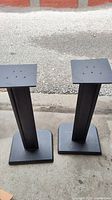 Pair of black metal speaker stands displayed on concrete floor with mounting holes visible on top plates. Paint rubbed off in some areas.