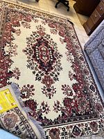 Full view of the cream-colored area rug with red, black, and beige floral medallion pattern and fringed edges on one side.