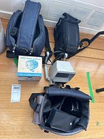 Four cameras and accessories on floor including camera bags, Super 8 Quartz Movie Lite projector box, and an Optex camera.