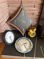 Four clocks displayed together on a floor corner: diamond-shaped Caravelle wall clock, black round wall clock, yellow twin bell alarm clock, and decorative round clock with floral border.