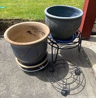 Two ceramic flowerpots on trays and three black metal plant stands on a concrete surface outdoors